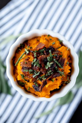 A closeup shot of a bowl of mashed sweet potato topped with fried bacon and greens on a striped cloth