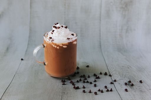 A closeup shot of a glass of hot chocolate on a table, with cream on top and chocolate pieces on the side