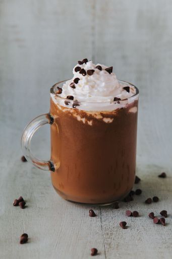 A vertical closeup of a glass of hot chocolate on a table, with cream on top and chocolate pieces on the side