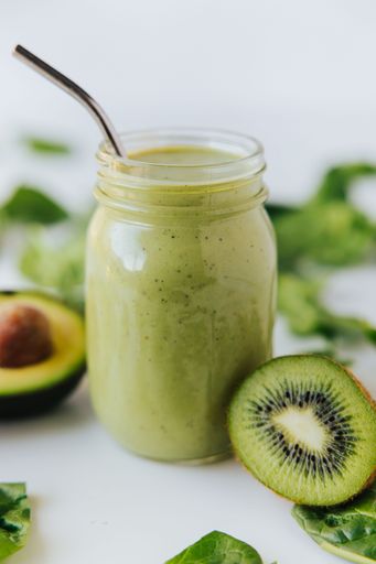 A macro shot of a jar with a healthy fresh green shake, with a half kiwi,a half avocado next to the glass,and fresh spinach leaves in the background