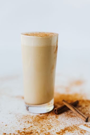 A closeup shot of a glass of a beige cinnamon mocha shake, sprinkled with cinnamon powder, surrounded with cinnamon powder and cinnamon sticks