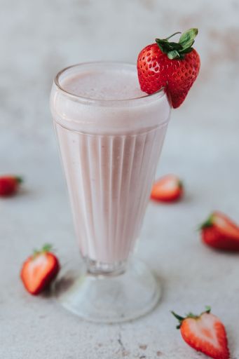 A closeup shot of a glass of strawberry milkshake, decorated with a fresh strawberry, and surrounded by blurred strawberries in the background