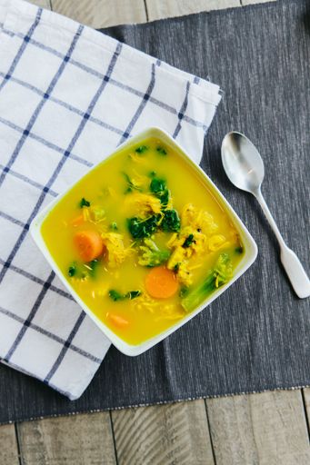A high angle of chicken vegetable soup inside a white plate with a spoon and handkerchief on the table