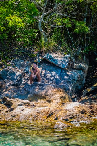 A vertical shot of an adult male sitting on the rocks near a river under the sunlight