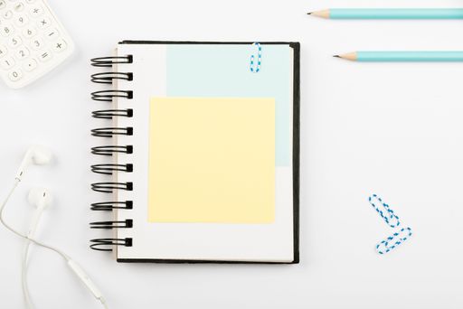Notebook With Message On Desk With Pens, Calculator, Headphones And Paperclips. Notepad With Information On Table With Pencils And Colored Clips.