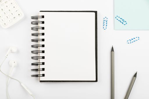 Notebook With Message On Desk With Pens, Calculator, Headphones And Paperclips. Notepad With Information On Table With Pencils And Colored Clips.