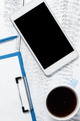 A vertical top view of a blank smartphone on a white table with a clipboard and a cup of coffee