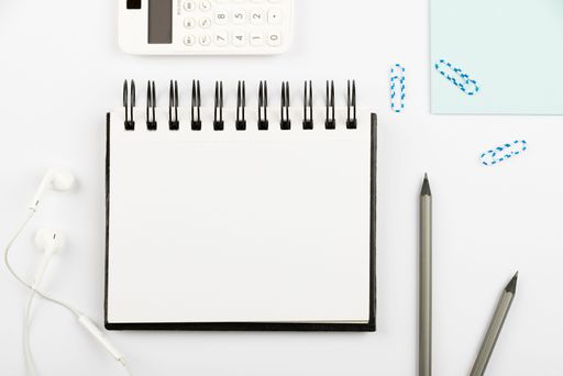 Notebook With Message On Desk With Pens, Calculator, Headphones And Paperclips. Notepad With Information On Table With Pencils And Colored Clips.