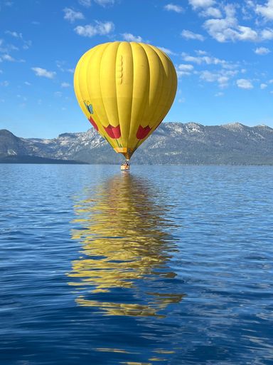 A vertical shot of a yellow hot air balloon flying over a tranquil lake against mountains