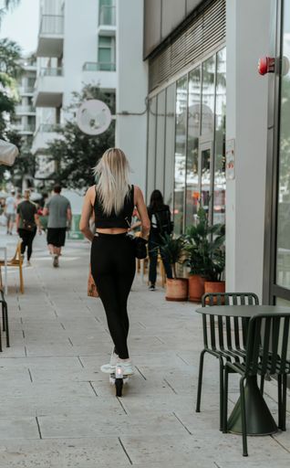 A vertical back view of a blond female in a black sports outfit riding on a scooter on the sidewalk crossing the shops