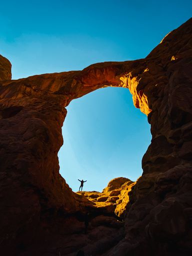 A vertical shot of a hiker in a stone arch against the background of blue sky. Arches National Park.