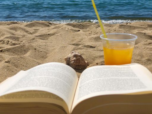 A selective focus shot of an opened book next to a cup of juice on a beach