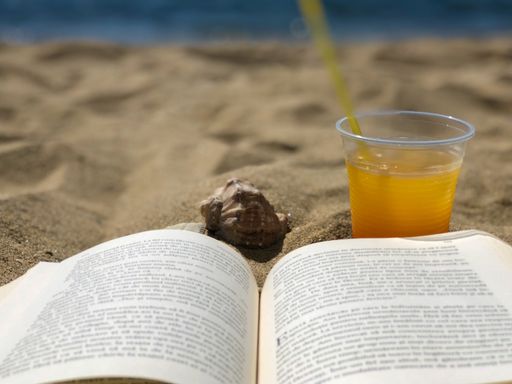 A selective focus shot of an opened book next to a cup of juice on a beach
