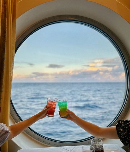 A vertical shot of two women's hands holding the glasses of cocktails against the ship's porthole