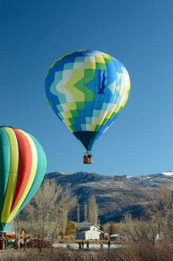 A vertical of a blue, yellow, green white patterned hot air balloon flying over the Colorado ridgeline against a clear, sunlit sky
