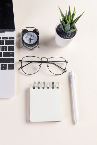 A vertical shot of an open laptop, glasses, a plant, a pen, a notepad, and an alarm clock