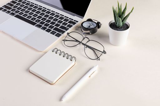A closeup shot of an open laptop, glasses, a plant, a pen, a notepad, and an alarm clock