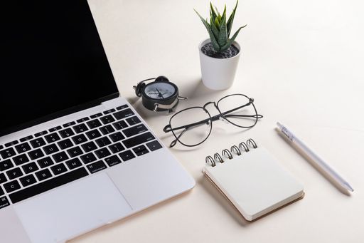 A closeup shot of an open laptop, glasses, a plant, a pen, a notepad, and an alarm clock