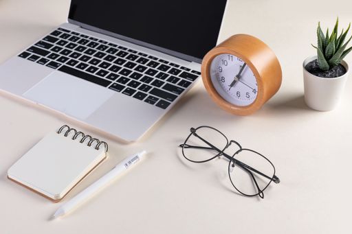 A closeup shot of an open laptop, glasses, notepad, pen, a clock, and a plant