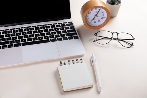 A closeup shot of an open laptop, glasses, notepad, pen, a clock, and a plant