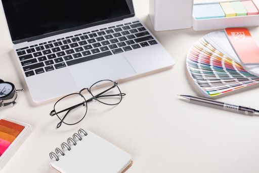 A closeup shot of an open laptop, glasses, a notepad, pen, and a fan ral
