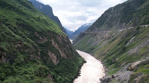 A bird's eye view shot of a wild long river in gorge between forested mountains and mountain road under cloudy sky