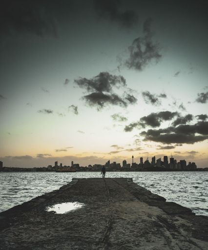 A vertical shot of a person stands on the edge of the road and looks at the sea with skyscrapers in the background