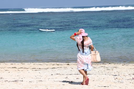 A female in a dress and pink hat walking on a sandy beach along the blue sea