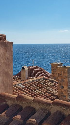 A vertical shot of the roof of a series of houses, with a cross on top of one, and the ocean in the background