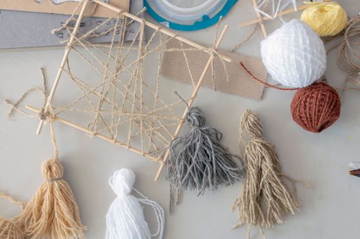 On table are materials and blanks (multi-colored woolen threads, wooden frame, tassels made of multi-colored thread) for creative activity of designer