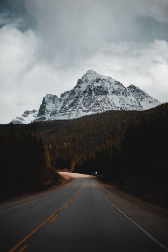 A vertical shot of long asphalt mountain road through forests leading to snowy rocky mountain