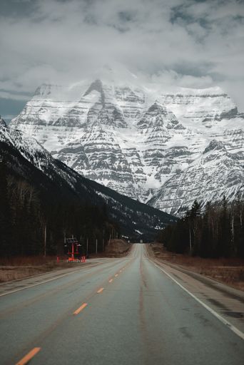 A vertical shot of long asphalt mountain road through forests leading to snowy rocky mountain