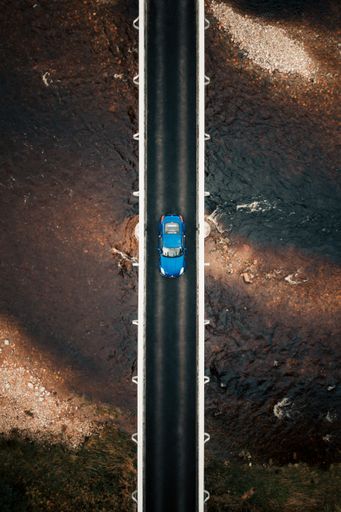 An aerial view of a blue car driving on a road bridge over a river