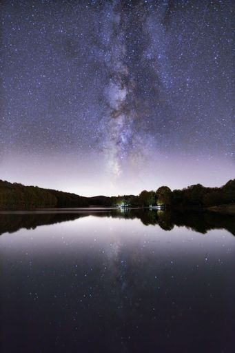 A vertical shot of a starry sky at night, with a cottage on the coast surrounded by silhouettes of trees, and their reflection on the lake surface