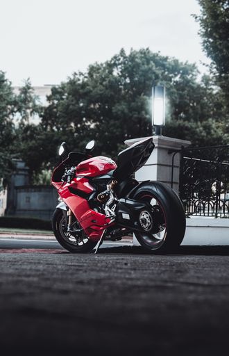 A side shot of a red motorcycle Ducati Panigale 1199, parked on the street, with a building and green trees in the background