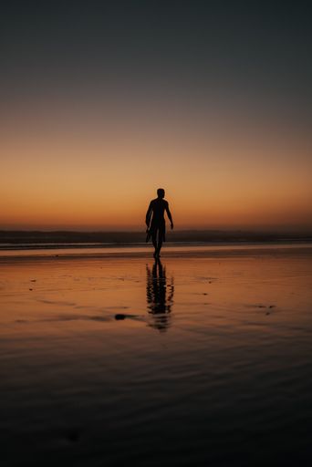 A vertical shot of a person with a surfing board walking on the beach at sunset perfect for wallpapers