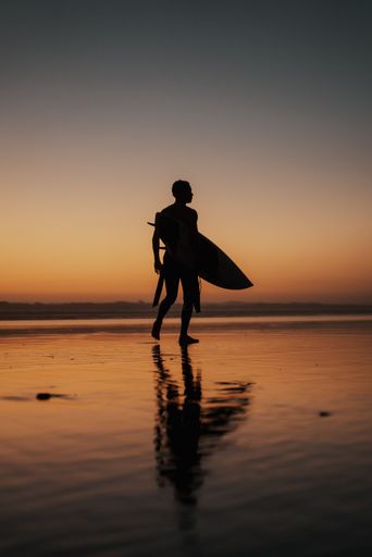 A vertical shot of a person with a surfing board walking on the beach at sunset perfect for wallpapers