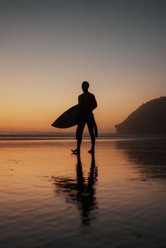 A vertical shot of a person with a surfing board walking on the beach at sunset perfect for wallpapers