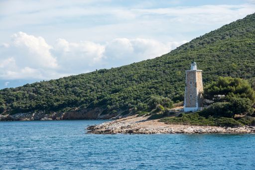 A beautiful scene of the small lighthouse by the sea surrounded by green hills in the daytime in Trikeri, Greece