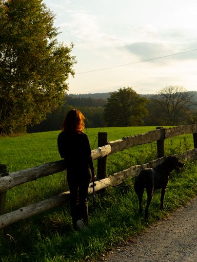 A woman and a black labrador retriever companion standing in front of a wooden fence in grassy meadow