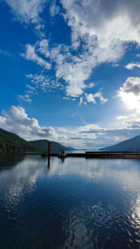 A vertical shot of a pier on a tranquil lake reflecting a cloudy blue sky surrounded by green hills