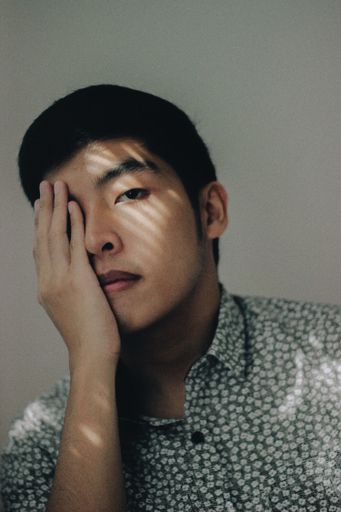 A vertical shot of an Asian male wearing a button-up shirt covering his eye with his hand