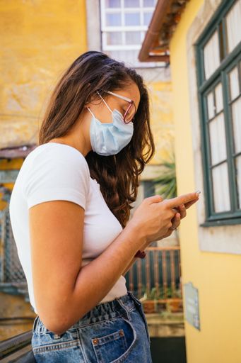 A shallow focus shot of a young beautiful Hispanic woman wearing a protective mask while using her mobile phone outdoors