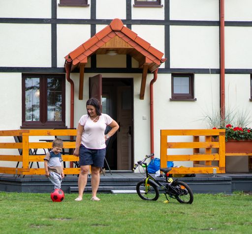 Woman standing next to young child with a ball next to small bicycle in front of a vacation house entrance on a warm summer day.