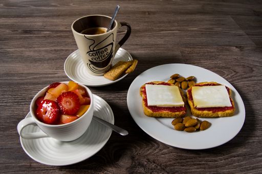 A closeup of a cup of coffee on the table with fruits, bread slices with jam, and almonds on it