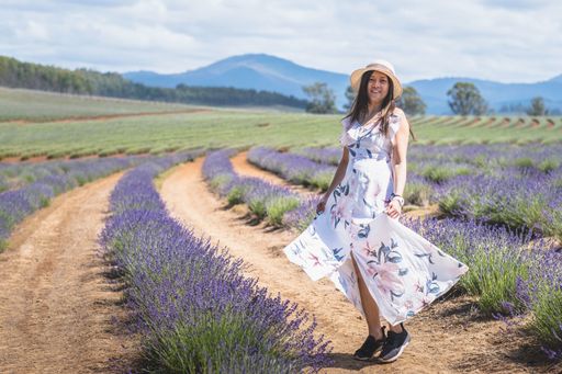 Asian Chinese mature woman in stylish dress looking away and walking amidst bushes of fresh lavender in summer cloudy day