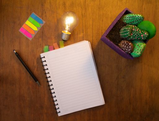 A flat lay shot of a copybook, a pen, a lamp, colorful stickers and decorative artificial cactus in a pot