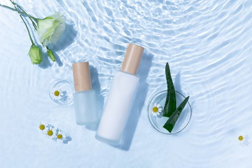 A top view of skincare bottles on a light blue water surface with aloe vera and daisy flowers