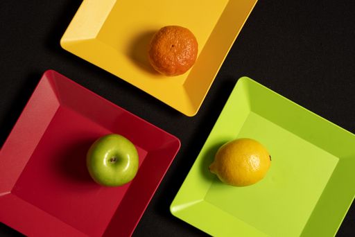 A top view of an apple, a lemon, and a tangerine on different colored plates, all three placed on a black surface