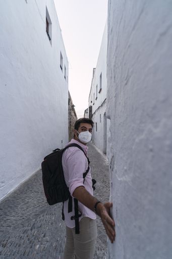 A young male with a backpack wearing a facemask and walking through an alley in Arcos de la Frontera, Spain
